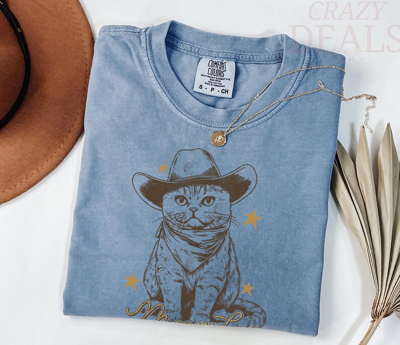 May include: A light blue t-shirt featuring a graphic of a cat wearing a cowboy hat and bandana. The cat is surrounded by gold stars and a rope design. The shirt has a Comfort Colors tag.