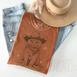 May include: Rust-colored t-shirt featuring a cat wearing a cowboy hat and bandana, with the word "Meowdy" in gold script. The shirt is paired with light-wash jeans, a tan hat, and a gold necklace. The words "Crazy Deals" are visible in the bottom right corner.