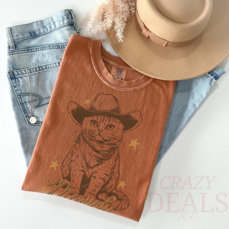 May include: Rust-colored t-shirt featuring a cat wearing a cowboy hat and bandana, with the word "Meowdy" in gold script. The shirt is paired with light-wash jeans, a tan hat, and a gold necklace. The words "Crazy Deals" are visible in the bottom right corner.