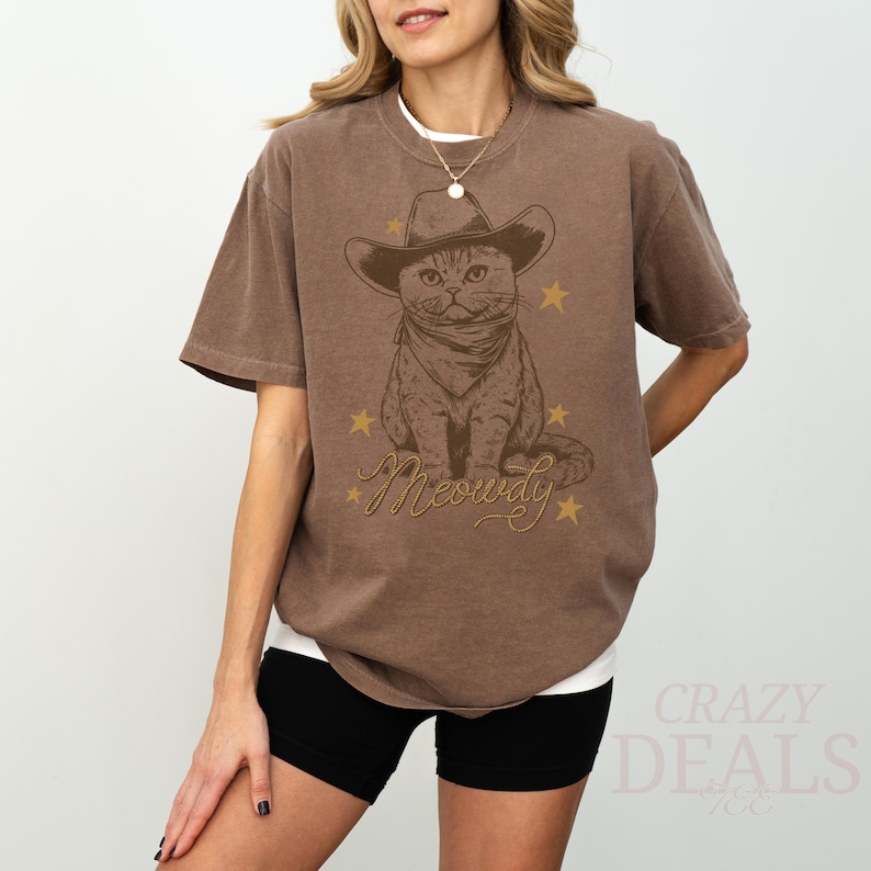 May include: Brown t-shirt featuring a cat wearing a cowboy hat and bandana, with the word "Meowdy" in a decorative font. Gold stars surround the cat graphic. The shirt is a relaxed fit.