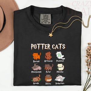 Pu&ograve; includere: T-shirt nera con la scritta "POTTER CATS" e illustrazioni di gatti dei cartoni animati. La maglietta presenta vari personaggi di gatti con nomi come "Rawrnald" e "Hermeownie". Una collana con ciondolo a stella dorata &egrave; drappeggiata sulla maglietta.