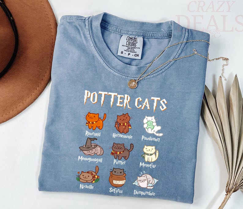 Pu&ograve; includere: Una maglietta blu Comfort Colors con la scritta "POTTER CATS" e illustrazioni di gatti dei cartoni animati. La maglietta ha un girocollo e maniche corte. Una collana d'oro con un ciondolo &egrave; drappeggiata sulla maglietta.