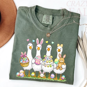 Peut inclure: T-shirt vert olive avec un motif de Pâques fantaisiste. Le graphisme représente quatre oies de dessin animé ornées d'œufs de Pâques, d'oreilles de lapin et d'un lapin en peluche. Un collier doré est drapé sur le t-shirt. Le texte "CRAZY DEALS" est visible en arrière-plan.