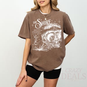 May include: Brown t-shirt with the text "THE SHIRE" and a graphic of a book, pipe, and moon. The shirt also has the quote "All you have to decide is what to do with the time that is given to us."
