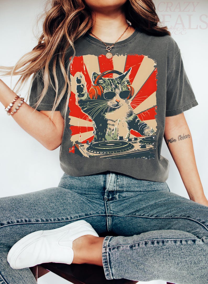 May include: Dark grey t-shirt with a cat DJ graphic wearing sunglasses and headphones, set against a red and beige sunburst. The cat is behind a turntable. The text "CRAZY DEALS CO" is visible at the top of the t-shirt.