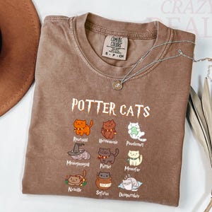 Pu&ograve; includere: Una maglietta marrone Comfort Colors con la scritta "POTTER CATS" e illustrazioni di gatti dei cartoni animati. La maglietta ha un girocollo e maniche corte. Una collana d'argento con un piccolo ciondolo &egrave; drappeggiata sulla maglietta.