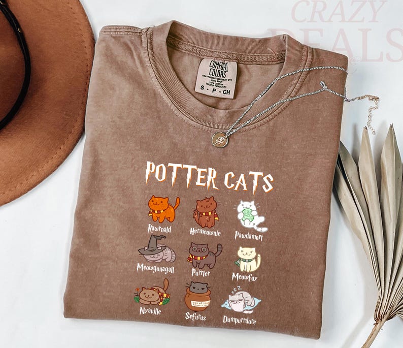 Pu&ograve; includere: Una maglietta marrone Comfort Colors con la scritta "POTTER CATS" e illustrazioni di gatti dei cartoni animati. La maglietta ha un girocollo e maniche corte. Una collana d'argento con un piccolo ciondolo &egrave; drappeggiata sulla maglietta.