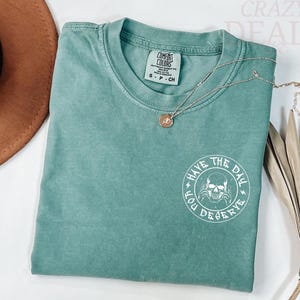Puede incluir: Una camiseta Comfort Colors de color verde azulado con cuello redondo y mangas cortas. La camiseta presenta un gr&aacute;fico blanco de una calavera con el texto "HAVE THE DAY YOU DESERVE". Un collar dorado con un colgante descansa sobre la camiseta.