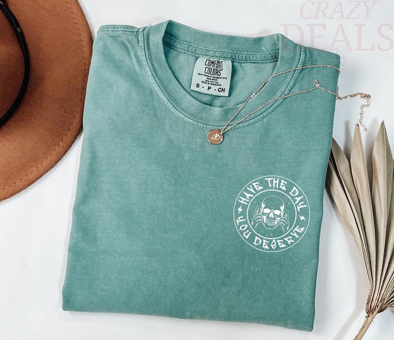 Puede incluir: Una camiseta Comfort Colors de color verde azulado con cuello redondo y mangas cortas. La camiseta presenta un gr&aacute;fico blanco de una calavera con el texto "HAVE THE DAY YOU DESERVE". Un collar dorado con un colgante descansa sobre la camiseta.