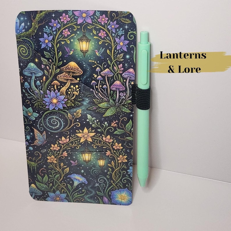 May include: A rectangular notebook cover featuring a dark, enchanted forest print with glowing lanterns, purple and blue mushrooms, flowers, and butterflies. A light mint green retractable pen is held in a black elastic loop on the side.