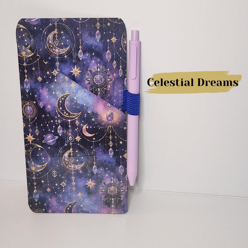 May include: A rectangular notebook with a dark purple and blue celestial pattern featuring gold moons, stars, planets, and hanging crystals. A light purple pen is held in place by a blue elastic loop on the side. The text Celestial Dreams is visible.