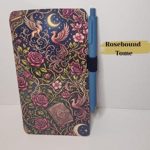 May include: A rectangular notebook cover featuring a dark background with pink roses, golden vines, a crescent moon, and phoenix illustrations. A small book icon is printed on the cover. A blue pen is attached to the side with a black elastic loop.