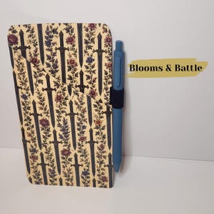 May include: A rectangular notebook with a cream-colored cover featuring a repeating pattern of dark gray swords and colorful floral vines. A blue retractable pen is held in a black elastic loop on the side. The text Blooms & Battle is visible.