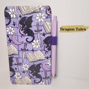May include: A rectangular notebook with a purple grid background featuring a repeating pattern of black dragons, open books, daggers, white flowers, and purple crescent moons. A light purple pen is held by an elastic loop on the side. Text reads Dragon Tales.