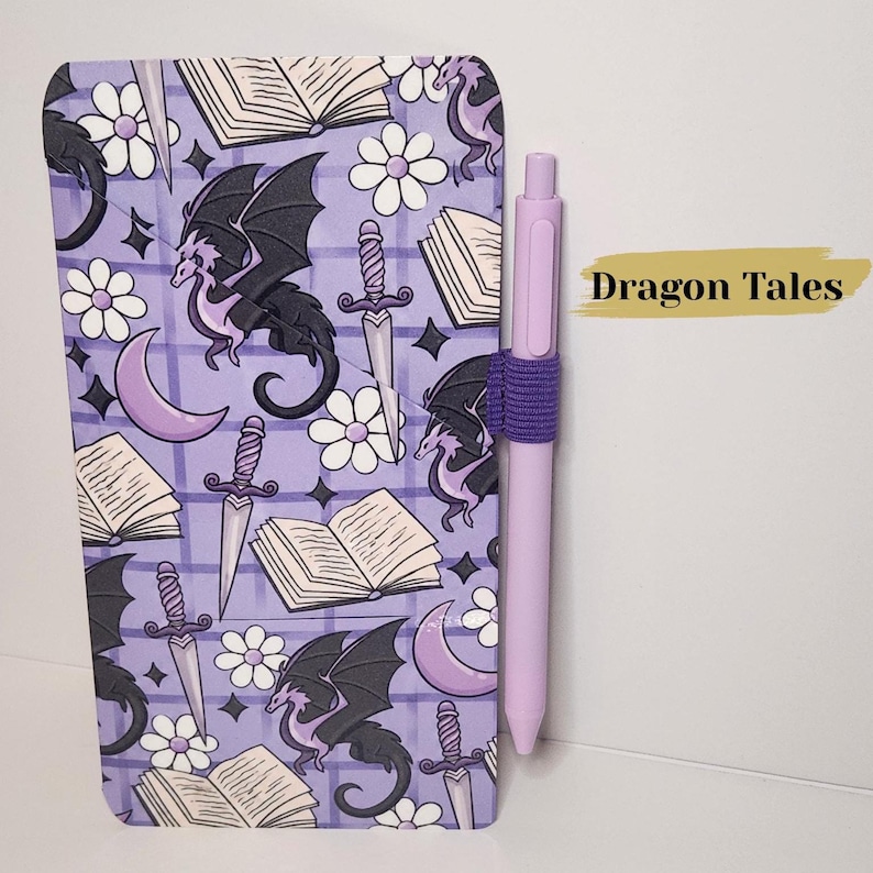 May include: A rectangular notebook with a purple grid background featuring a repeating pattern of black dragons, open books, daggers, white flowers, and purple crescent moons. A light purple pen is held by an elastic loop on the side. Text reads Dragon Tales.