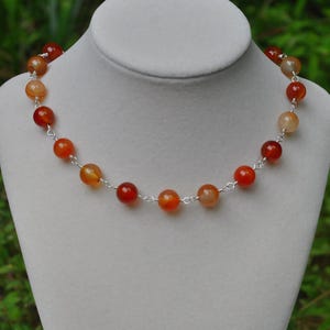 May include: A necklace featuring round, polished carnelian beads in shades of orange and deep red, connected by thin silver-toned metal links. The beads are arranged in a single strand, showcasing a smooth, translucent texture and varied warm tones.