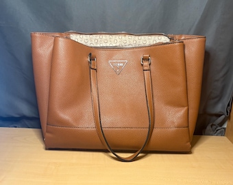 May include: A brown leather tote bag with a zip closure and two handles. The bag has a textured surface and a silver triangle logo on the front. The interior lining features a repeating pattern of the brand's logo.