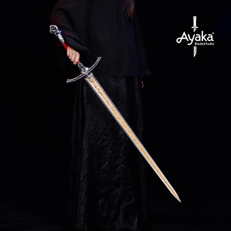 May include: A long, silver sword with a gold blade and ornate hilt is held against a black background. The hilt features a red grip and intricate detailing. The blade has gold inscriptions. The logo "Ayaka BladeStudio" is in the upper right corner.