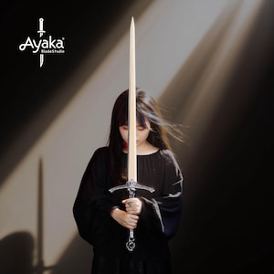 May include: A long sword with a light-colored blade and ornate silver hilt is held by a person wearing a black top. The sword's blade is centered in the frame, with the hilt at the bottom. The logo "Ayaka BladeStudio" is in the upper left corner.