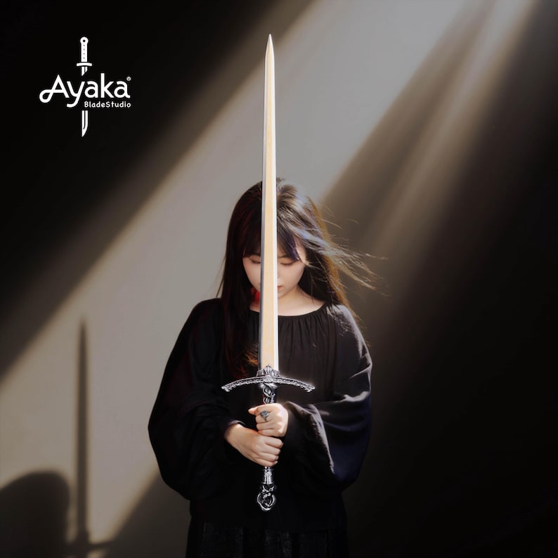May include: A long sword with a light-colored blade and ornate silver hilt is held by a person wearing a black top. The sword's blade is centered in the frame, with the hilt at the bottom. The logo "Ayaka BladeStudio" is in the upper left corner.