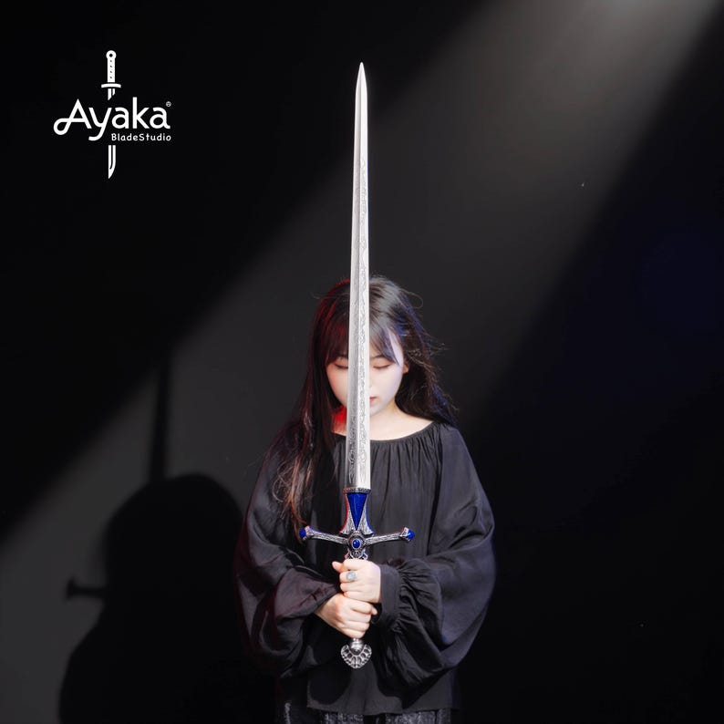 May include: A long, silver sword with a blue and silver hilt is held vertically. The sword's blade is centered in the frame, with a person holding the sword. The logo "Ayaka BladeStudio" is in the upper left corner.