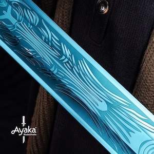 May include: Close-up of a blue sword blade with intricate etched designs. The blade has a reflective surface and is held against a dark background. The logo "Ayaka BladeStudio" is visible.