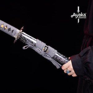 Op de afbeelding: Een close-up van een katana-zwaard. Het zwarte lemmet heeft een decoratief patroon en de handgreep is gewikkeld in grijze stof. Gouden accenten en een zilveren beschermkap zijn zichtbaar. De merknaam "Ayaka BladeStudio" is zichtbaar.