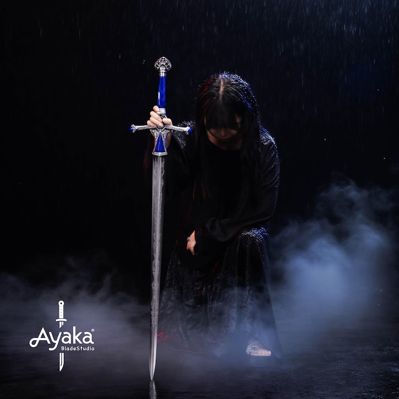 May include: A long, silver sword with a blue handle and ornate silver accents. The sword is held upright against a dark background with a person kneeling in a black robe. The image includes the text "Ayaka BladeStudio".