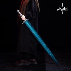 May include: A long, two-handed sword with a silver hilt and a vibrant blue blade. The blade features a unique pattern and is held by a person wearing dark clothing. The Ayaka Blade Studio logo is visible in the upper right corner.