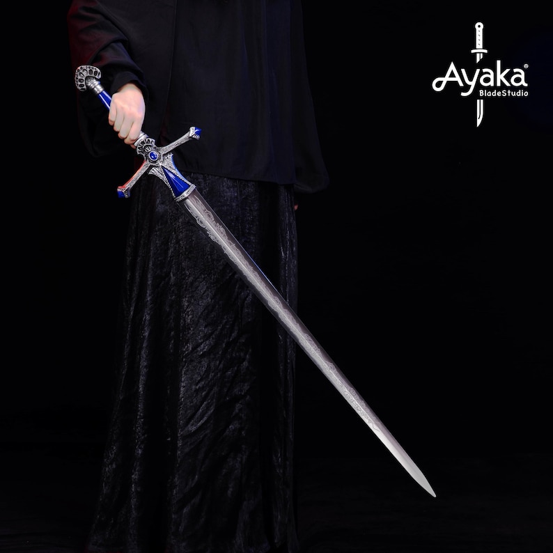 May include: A long, silver sword with a blue and silver hilt. The sword has a cross-shaped guard and a detailed design on the blade. The hilt features a blue gem and ornate silver accents. The sword is held against a dark background.