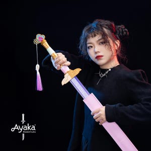 Könnte beinhalten: Ein dekoratives Schwert mit einem rosa-goldenen Griff, einer lila Klinge und einer rosa Scheide. Der Griff hat ein herzförmiges Design mit einer Quaste. Das Schwert stammt von Ayaka BladeStudio.