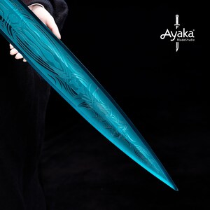 May include: A translucent, teal-colored sword with intricate etched designs. The blade has a pointed tip and is held against a black background. The logo "Ayaka BladeStudio" is visible in the upper right corner.