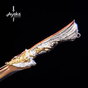 Könnte beinhalten: Verziertes Schwert mit silbernem und goldenem Griff, mit detailliertem Flügeldesign. Die Klinge hat einen kupferfarbenen Farbton. Der Schwertgriff hat ein Drachendesign. Das Logo "Ayaka BladeStudio" befindet sich oben links.