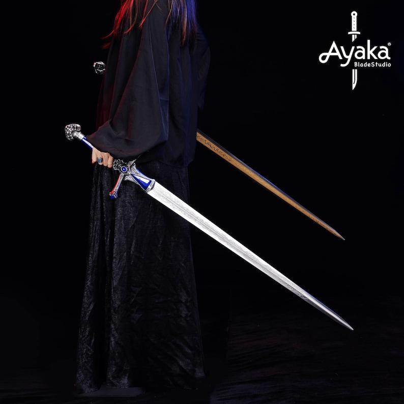 May include: A long, silver sword with a blue and silver hilt is held in front of a person wearing a black robe. A second sword with a wooden handle is visible in the background. The Ayaka BladeStudio logo is in the upper right corner.
