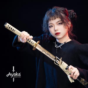 Puede incluir: Una katana con una hoja y empuñadura doradas, con un diseño de dragón. El mango está envuelto en un material color crema. La espada es sostenida por una persona con un suéter negro. El logotipo "Ayaka BladeStudio" está en la esquina inferior izquierda.
