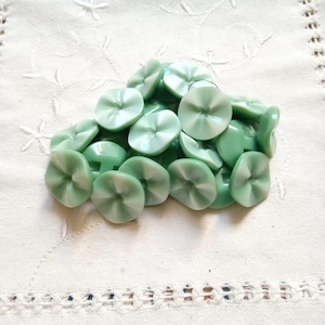 以下が含まれることがあります： 白い渦巻き模様の、薄緑色の花型のボタンの山。ボタンは光沢のある素材でできており、白い刺繍の生地の上に散らばっています。