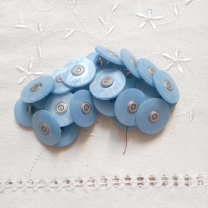 Könnte beinhalten: Eine Ansammlung hellblauer, runder Knöpfe mit silberfarbenen Zentren. Die Knöpfe sind auf einem weißen, bestickten Stoff angeordnet. Die Knöpfe haben einen perlmuttartigen Glanz und ein dekoratives Design in der Mitte.