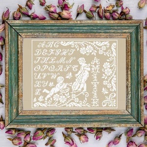 Peut inclure: Un cadre brodé au point de croix avec un fond beige et un cadre en bois vert et doré. L'échantillon présente des lettres brodées blanches, des motifs floraux et des figures. Des boutons de roses séchées entourent le cadre.