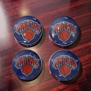 May include: Four round, blue coasters featuring the New York Knicks logo. Each coaster has the team name and a basketball in the center, with a white and gray border. The coasters are arranged in a square.
