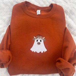 May include: A rust-colored crewneck sweatshirt featuring a central embroidered design of a Highland cow wearing a white ghost costume. The cow has brown horns, dark eyes, and a brown nose, with the ghost sheet draped over its body.