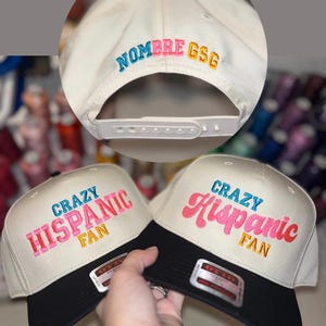 May include: Two beige and black baseball caps with the text "CRAZY HISPANIC FAN" embroidered in pink, blue, and gold. The back of the cap has the text "NOMBRE GSG" embroidered in blue, pink, and gold.