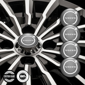 May include: Close-up of a black and silver car wheel with a Volvo logo center cap. The wheel has a glossy finish with white and black spokes. Four additional Volvo logo caps are displayed. The image also includes two circular custom made badges.