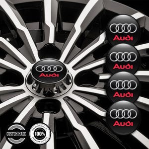 May include: Black and silver car wheel with white spokes. The center cap features the Audi logo in silver and the word "Audi" in red. Additional caps with the Audi logo are displayed. The image also includes "CUSTOM MADE" and "PREMIUM QUALITY" text.