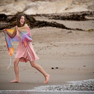 May include: A young person wearing a pink dress and a colorful shawl walks on a sandy beach with the ocean in the background.