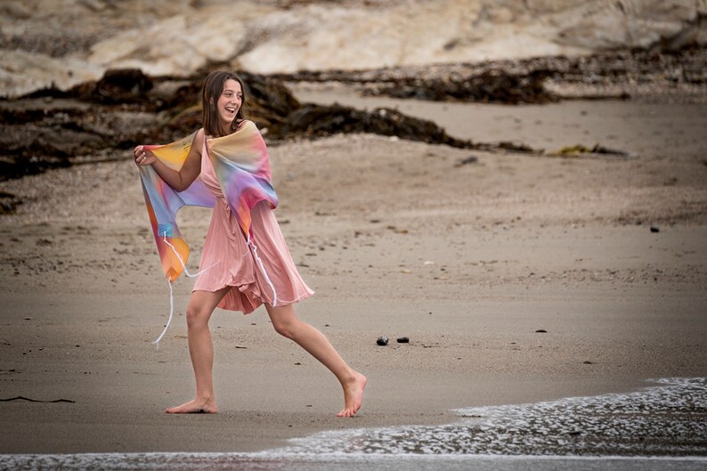 May include: A young person wearing a pink dress and a colorful shawl walks on a sandy beach with the ocean in the background.