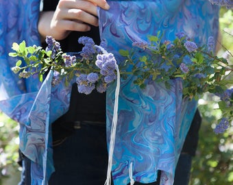 May include: A blue and purple patterned prayer shawl with white tassels. The shawl is draped over a person's shoulders and held in their hand. The shawl is surrounded by purple flowers.