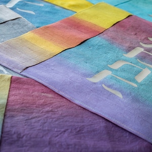 May include: A close-up of several colorful fabric squares with Hebrew lettering. The fabric is a gradient of colors, including purple, blue, yellow, and pink. The lettering is white and appears to be hand-painted.