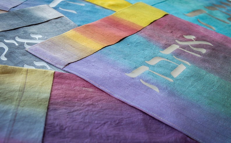 May include: A close-up of several colorful fabric squares with Hebrew lettering. The fabric is a gradient of colors, including purple, blue, yellow, and pink. The lettering is white and appears to be hand-painted.