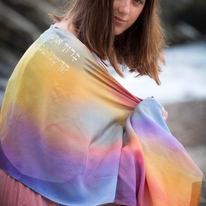 May include: A young person wearing a colorful ombre shawl with Hebrew text. The shawl is draped over their shoulders and is made of a soft, flowing fabric.