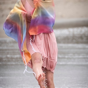 May include: A woman wearing a pink dress and a colorful shawl stands in shallow water on a beach. She is smiling and kicking her foot in the water, creating a splash.